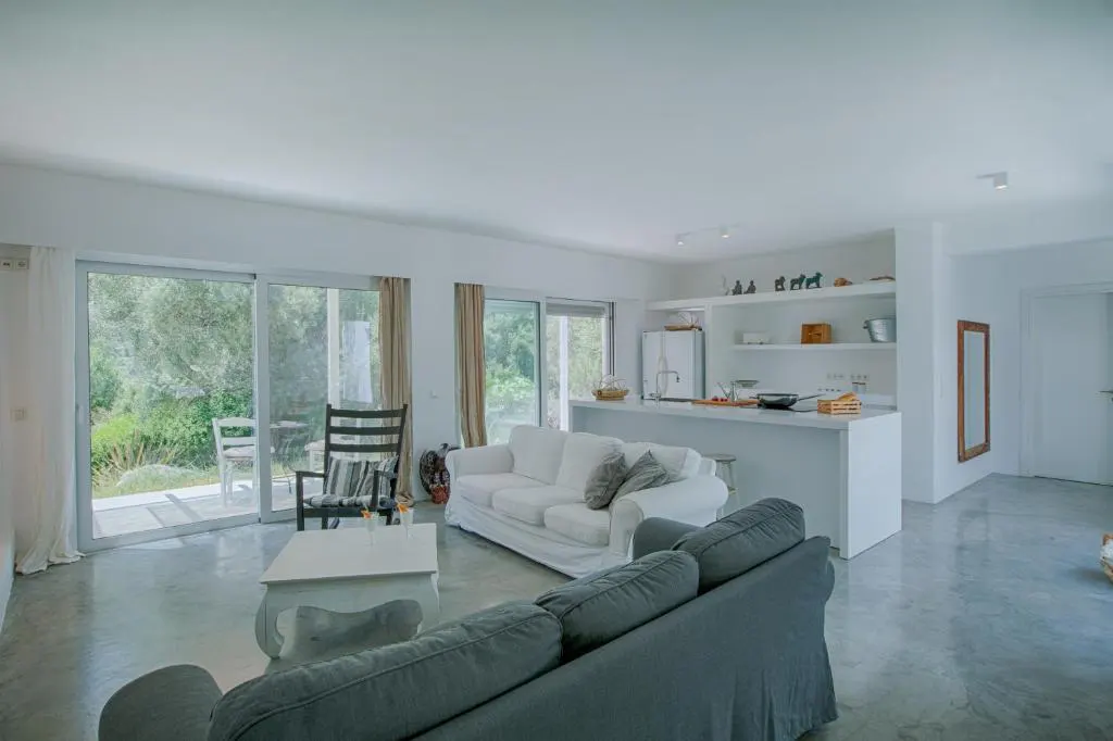 Open-concept living space with white walls, gray concrete floors, and large sliding glass doors to a green outdoor area.