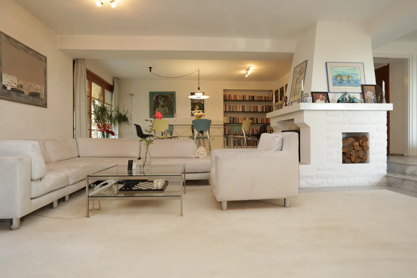 Bright, open living room with white sofa, chair, and fireplace. A glass coffee table holds a chess set. Bookshelves are visible in the background.