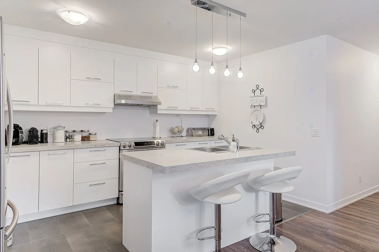 Bright, modern kitchen with white cabinets, island with two white bar stools, stainless steel appliances, and pendant lighting.