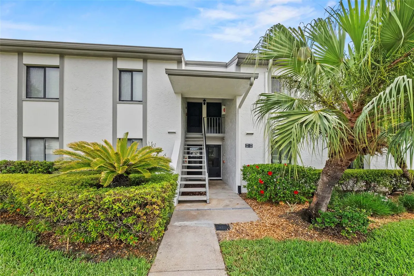 Exterior of a two-story white condo building with gray trim, stairs to the second floor, and lush landscaping.