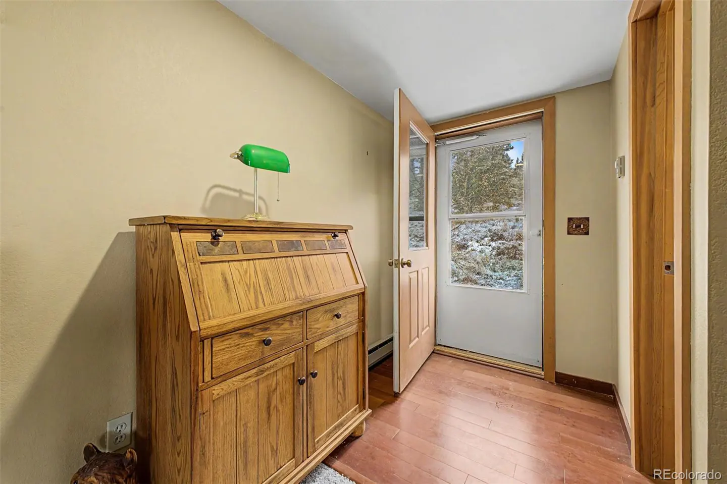 Entryway with a wooden desk topped with a green lamp, an open door with a snowy outdoor view, and hardwood floors.