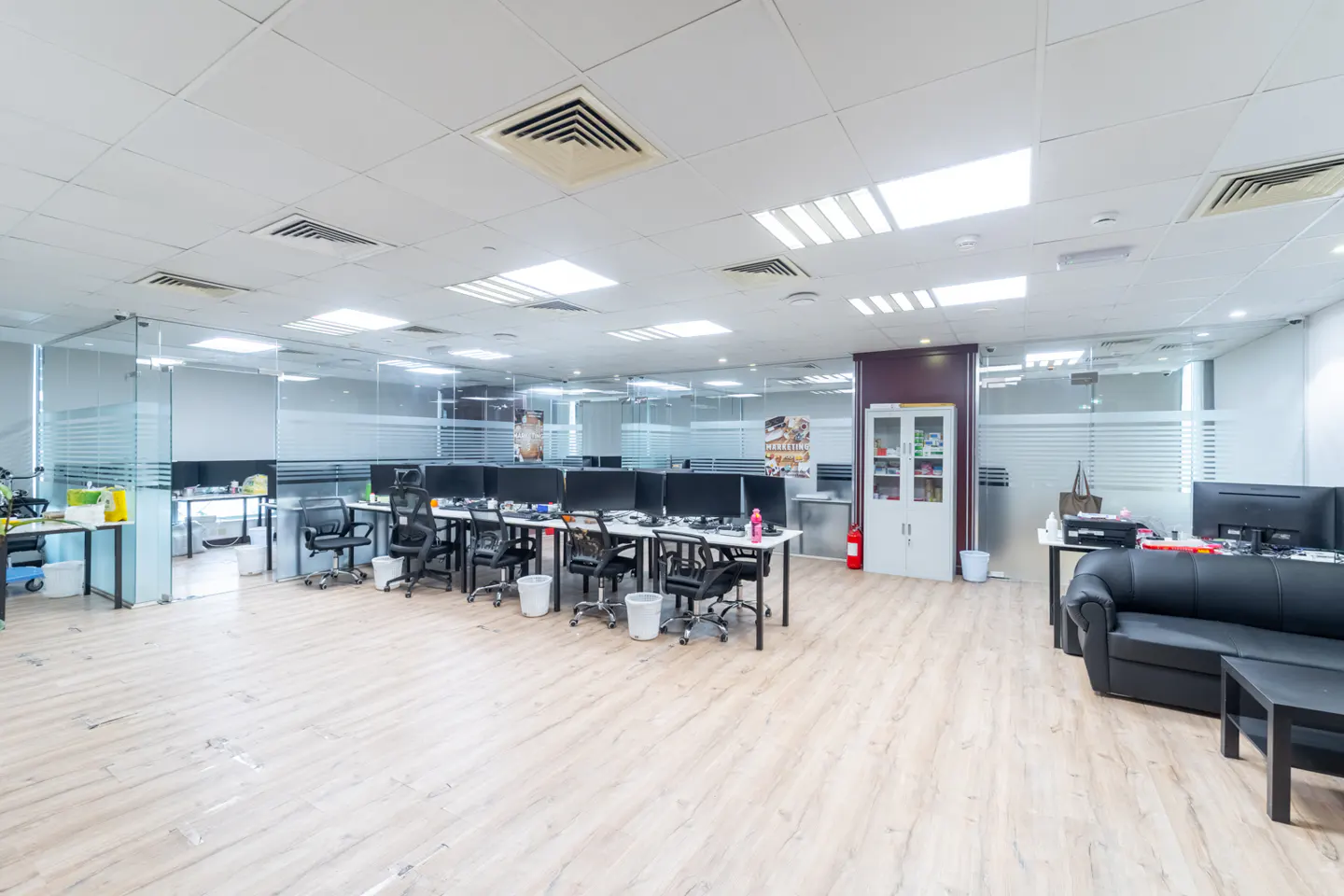 Bright, modern office space with rows of desks, black chairs, and computer monitors. Glass-walled offices and a black sofa add to the professional setting.