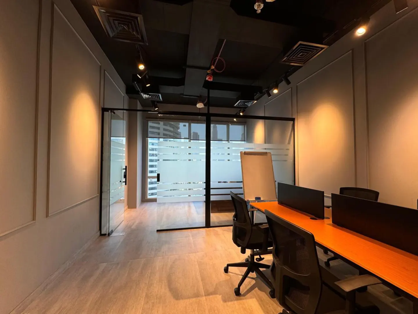 Office space with desks, black chairs, and frosted glass walls. A whiteboard stands near a window with a city view. The ceiling is dark with track lighting.
