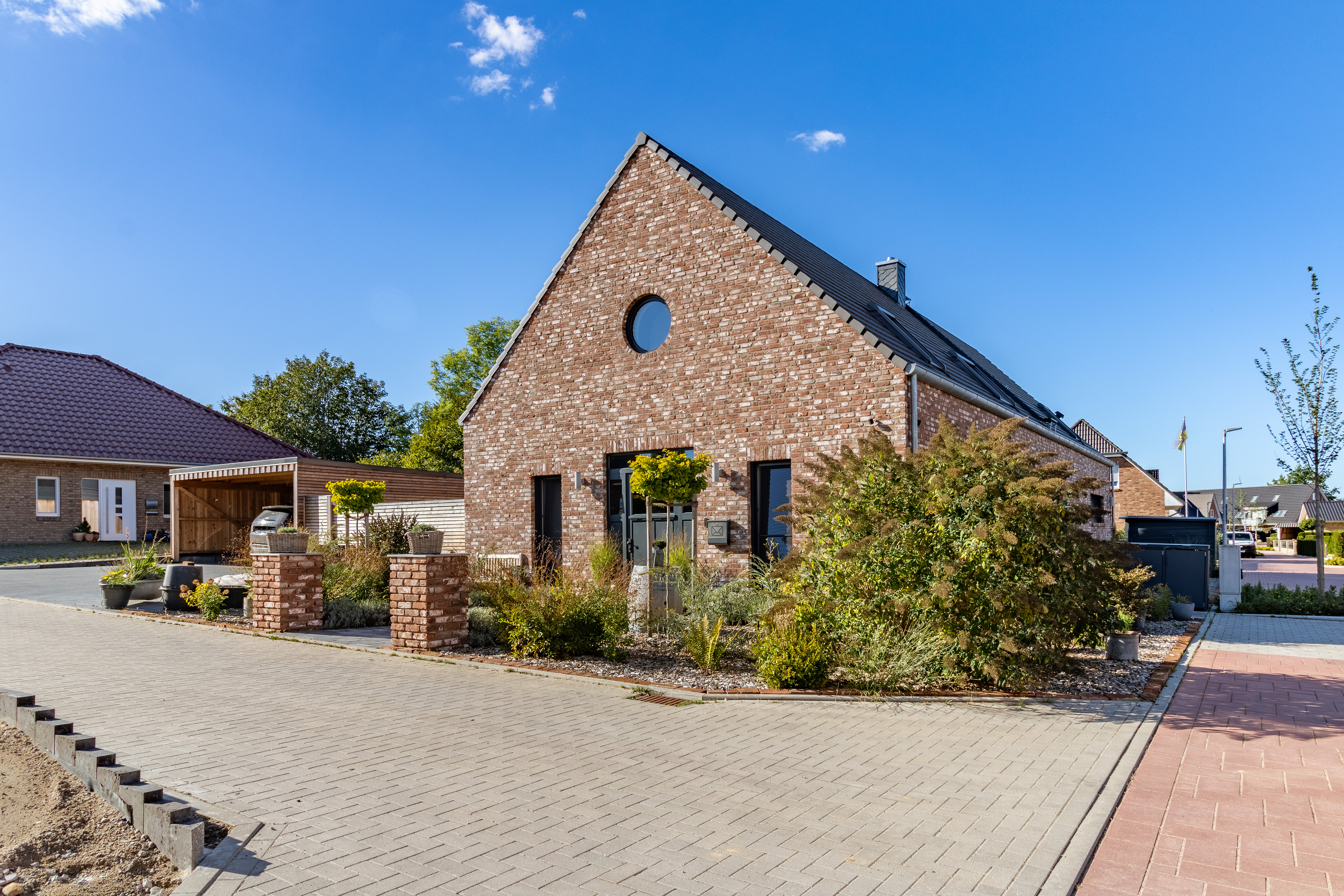 Modern brick house with a steep roof, circular window, and landscaped front yard under a clear blue sky. [AI-generated text]