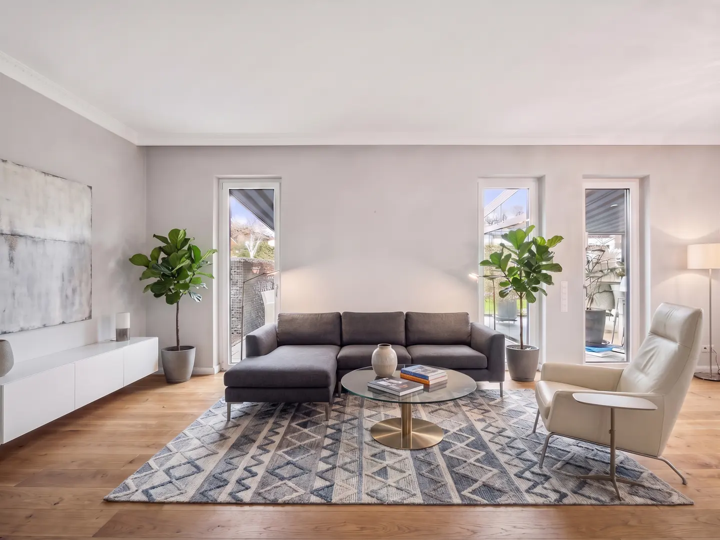 A modern living room with a gray sectional sofa, patterned rug, and light wood floors. Three windows provide natural light.