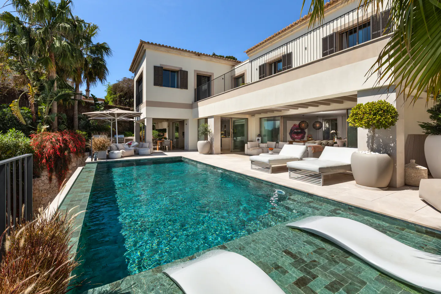 Luxury villa with a turquoise pool, white lounge chairs, and lush greenery under a clear blue sky.