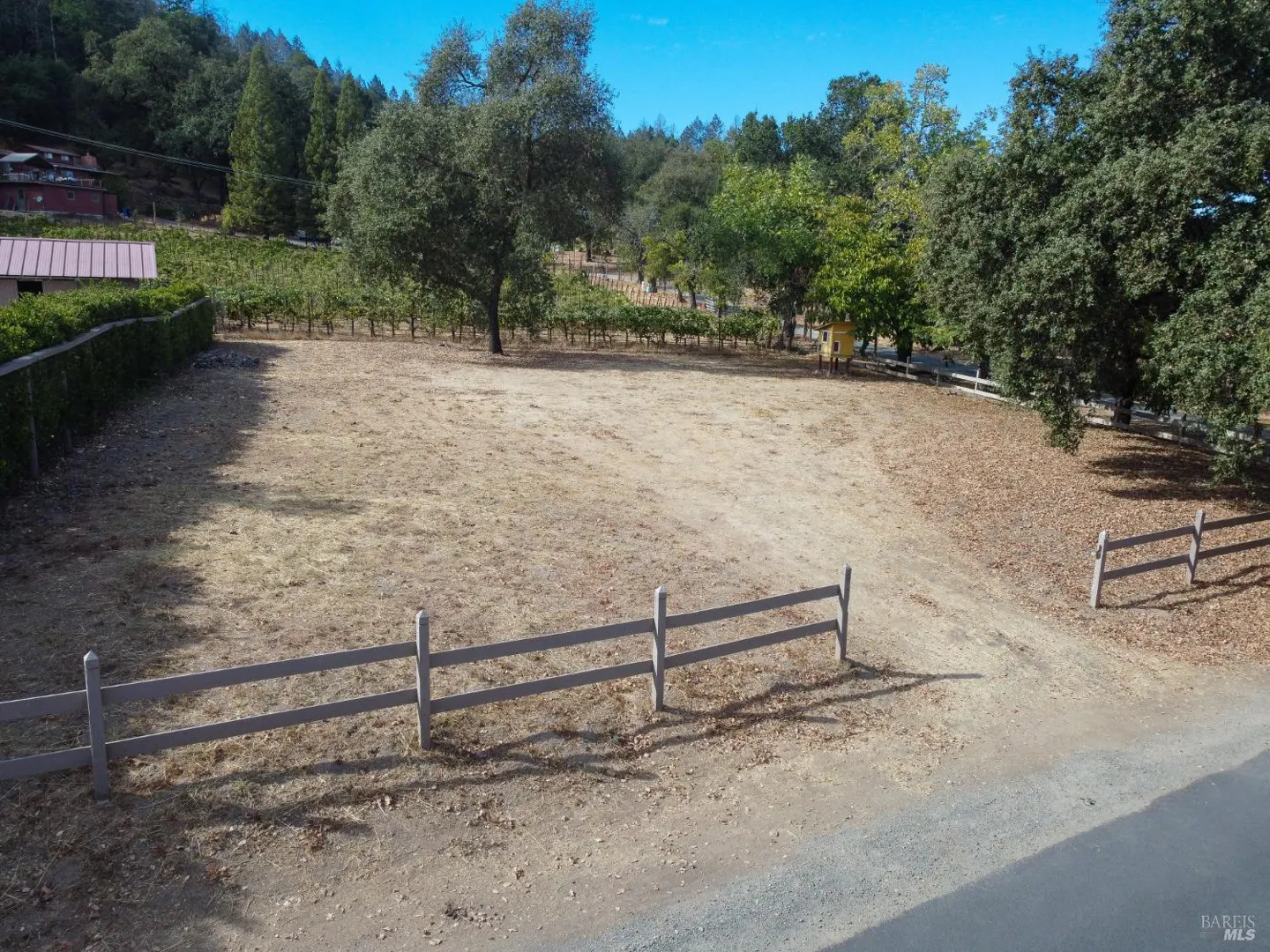 A vacant lot with a wooden fence, dry grass, and trees in the background. A vineyard is visible behind the lot.