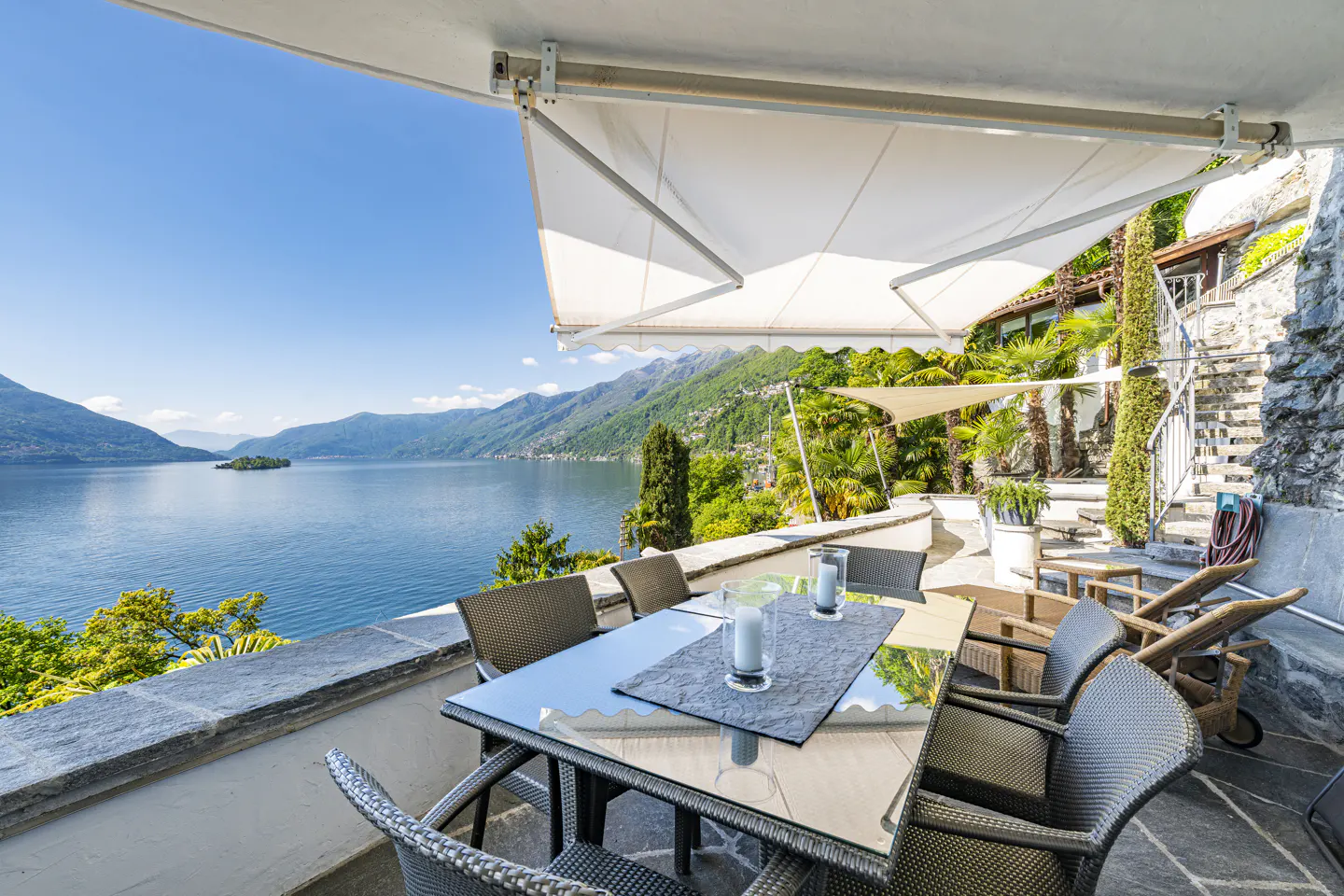 Outdoor patio with a glass table, wicker chairs, and a view of a blue lake and mountains under a sunny sky.