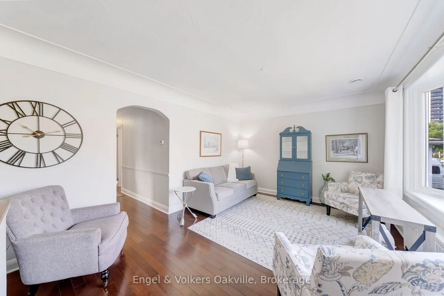 Bright living room with hardwood floors, a gray sofa, blue hutch, and floral armchairs. A large clock hangs on the wall.