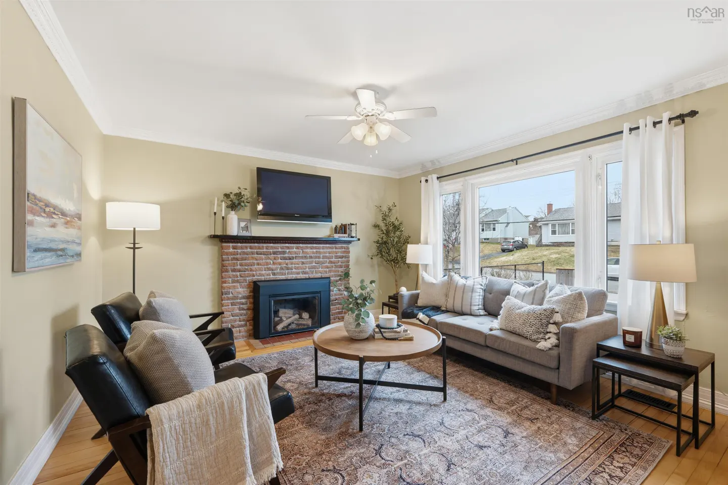 Living room with a brick fireplace, TV, gray sofa, wood floor, and a large window with white curtains.