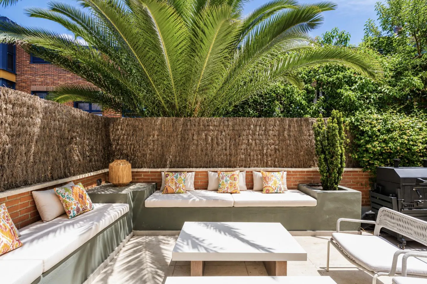 A sunny outdoor patio with built-in concrete benches, white cushions, colorful patterned pillows, and a central table.