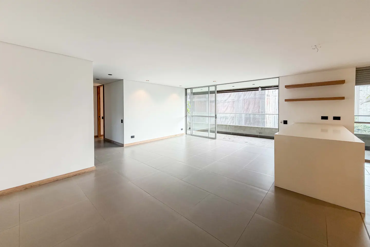 Bright, empty room with white walls, gray tile floor, and sliding glass doors to a balcony. A white kitchen island and wood shelves are on the right.
