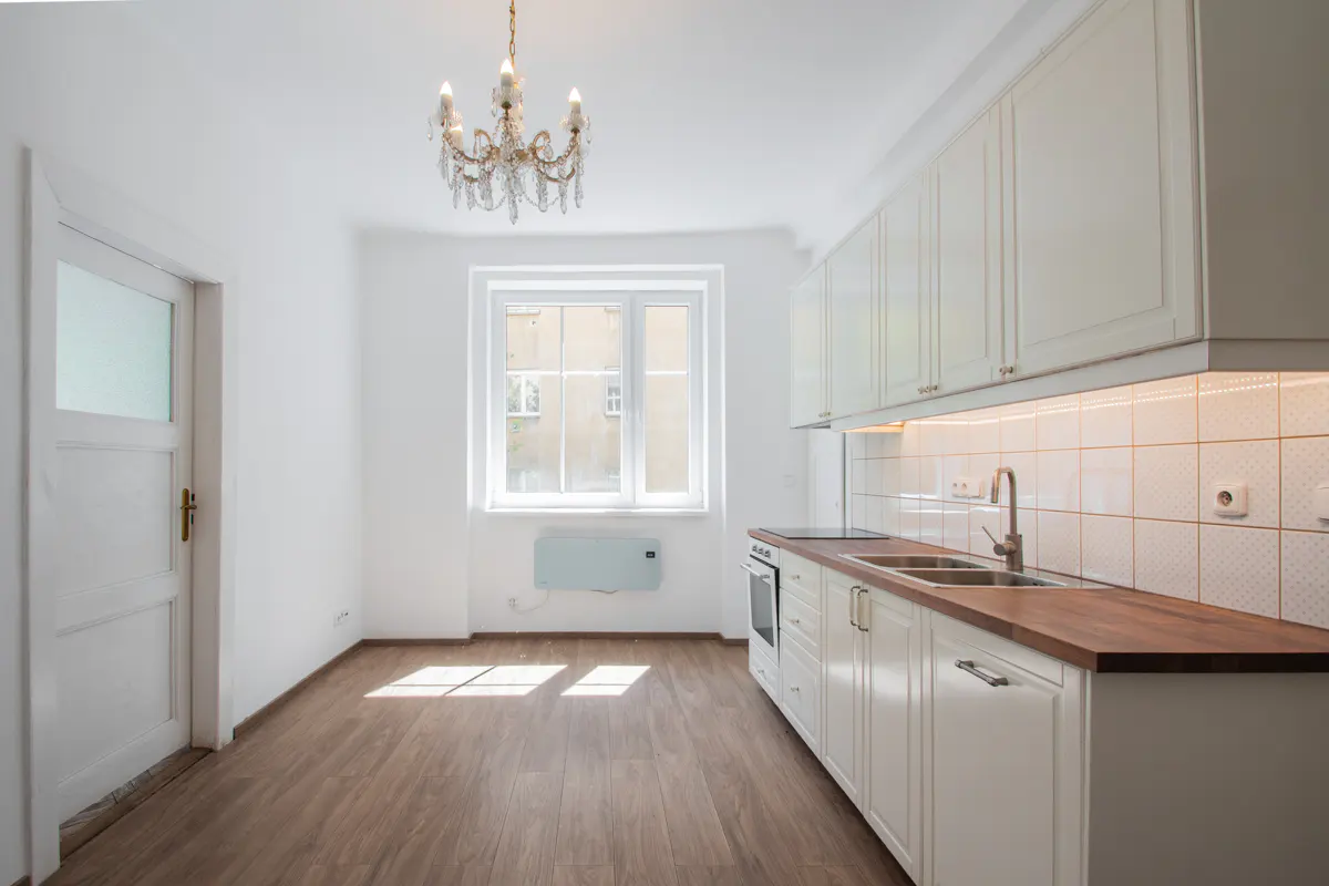 Bright kitchen with white cabinets, wood countertops, and a chandelier. A window lets in natural light, illuminating the wood floor.