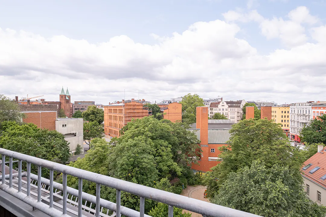 Cityscape view from a balcony with a gray railing. Buildings are brick and yellow, with green trees and a cloudy sky.