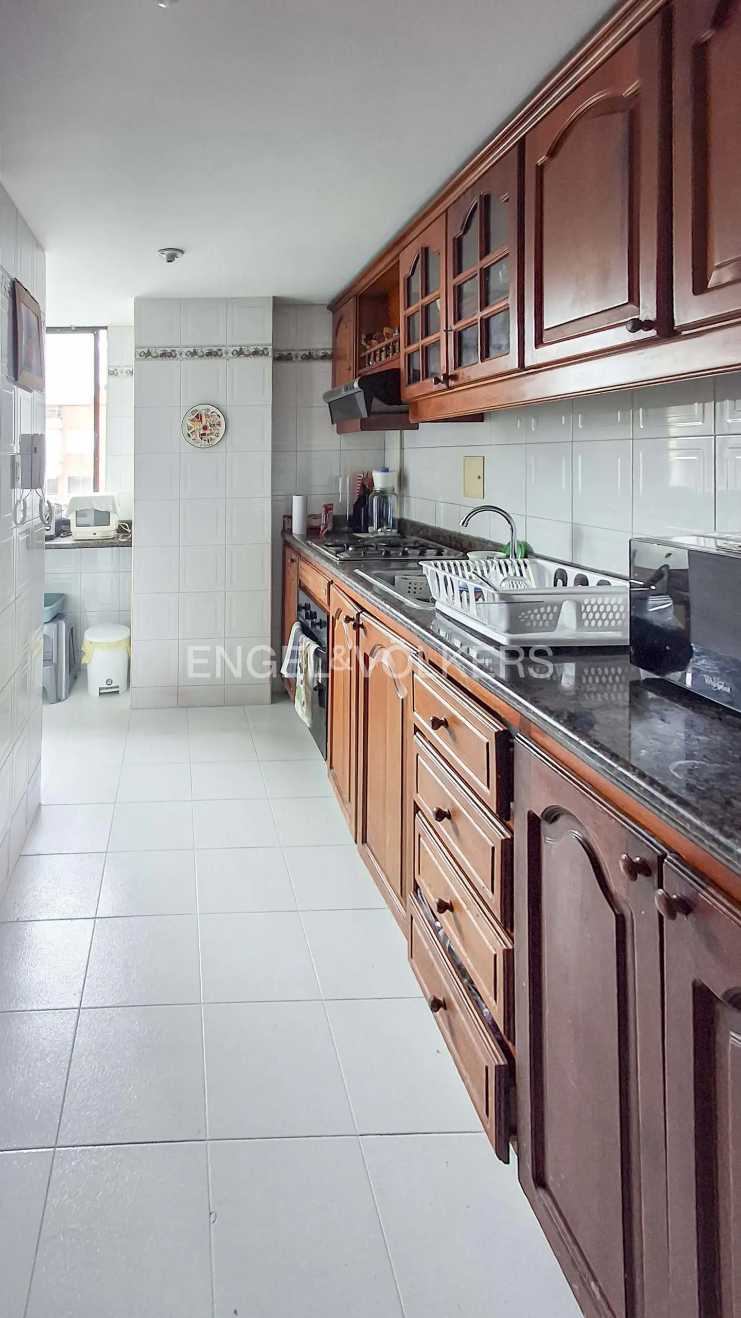A kitchen with white tile floors and walls, and dark wood cabinets. A window is visible in the background.