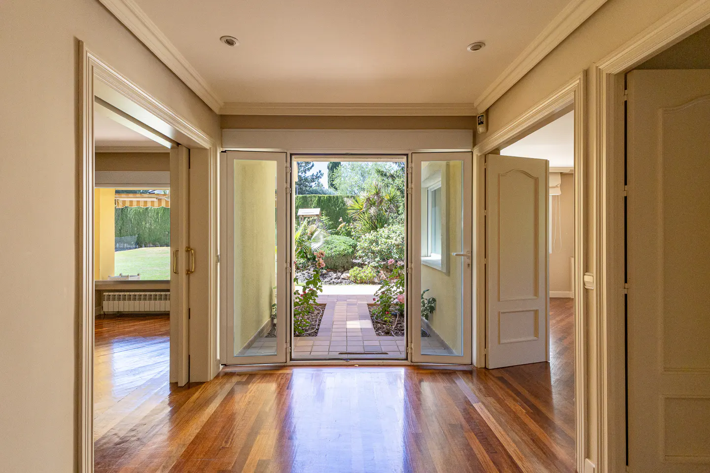 Bright hallway with polished wood floors leading to open glass doors revealing a lush, sunny garden patio.