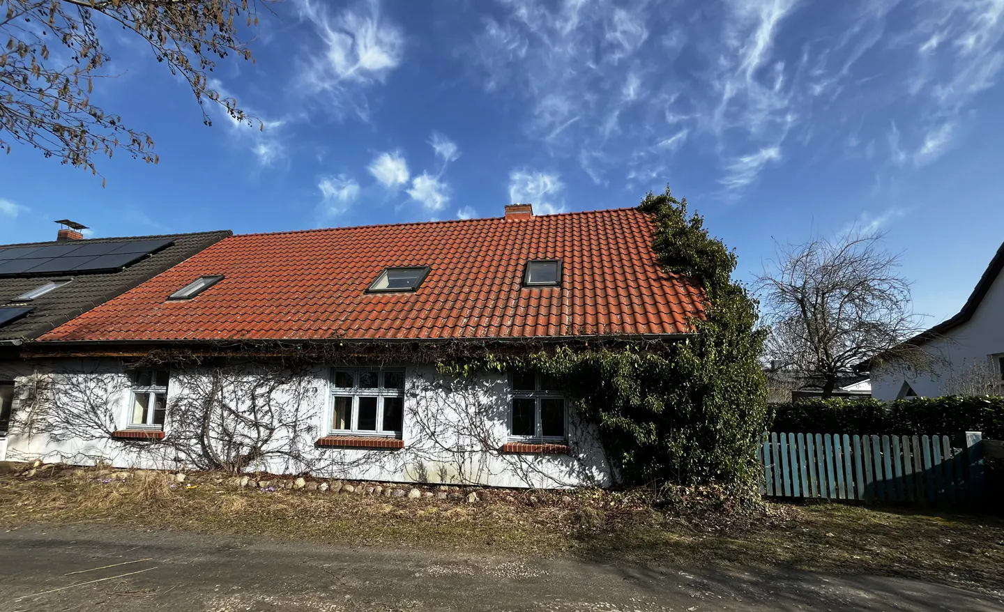 Exterior view of a white house with a red tile roof, dormer windows, and climbing vines against a blue sky.