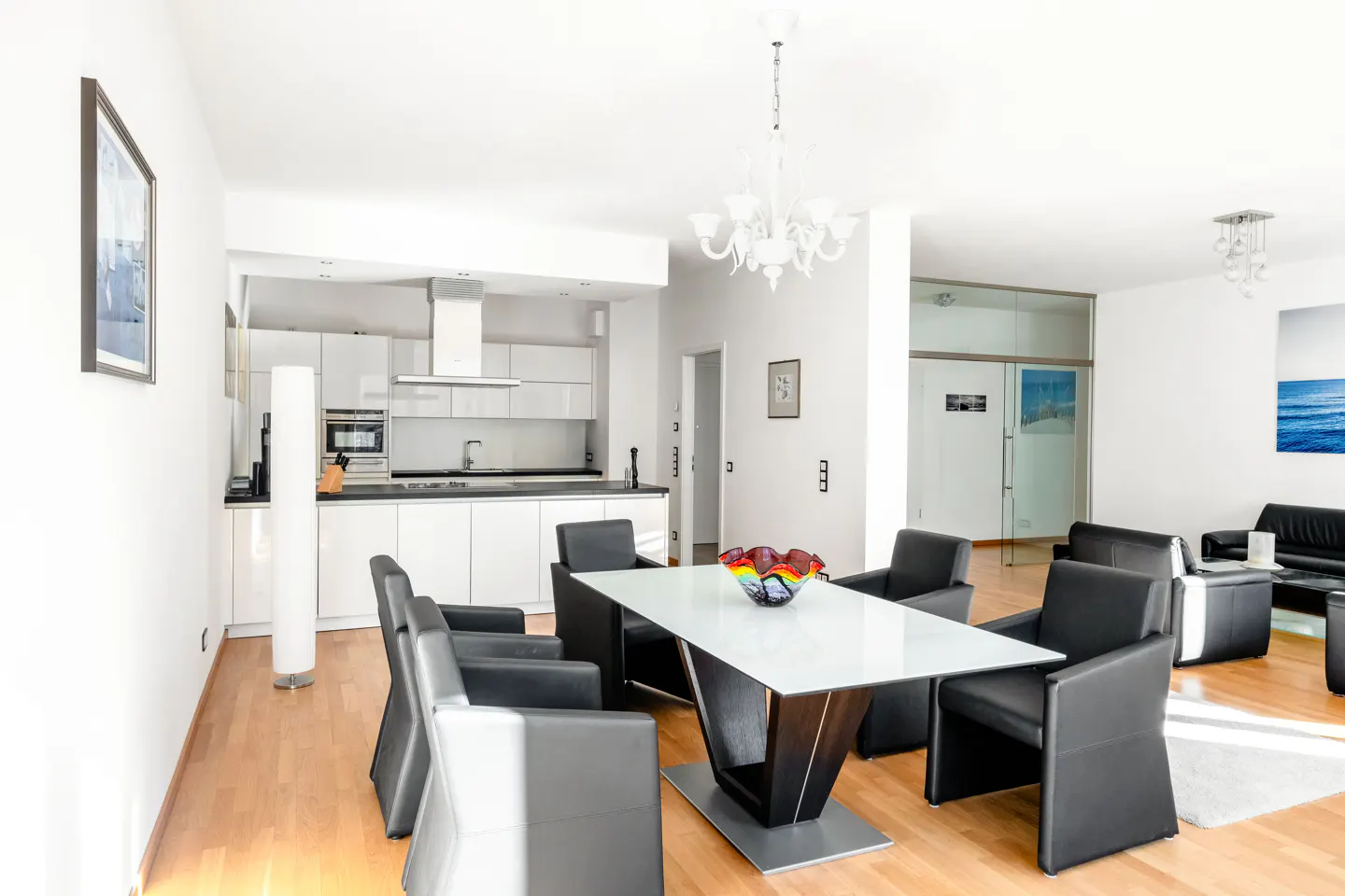 Bright, modern open-concept living space with white walls, wood floors, and black leather chairs around a white dining table. Kitchen in background.