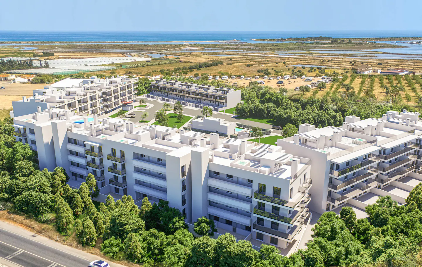 Aerial view of modern white apartment buildings with balconies, surrounded by green trees, with a distant view of the ocean.