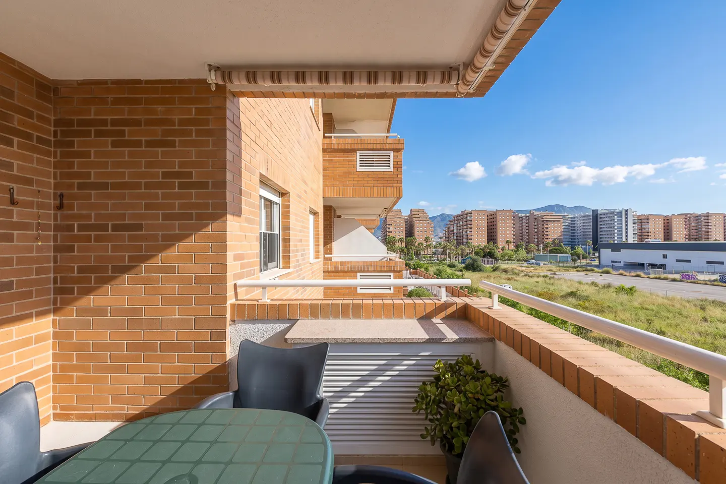 Balcony view of a brick building with a green table, chairs, and a plant. City buildings are visible in the background under a blue sky.