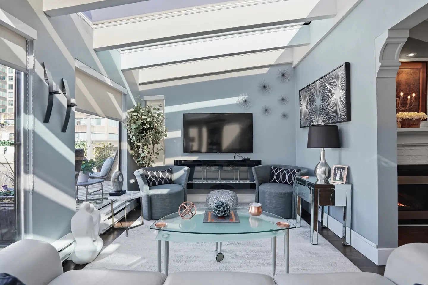 Bright living room with skylights, blue walls, and gray furniture. A glass coffee table sits on a white rug. A TV hangs above a black console.