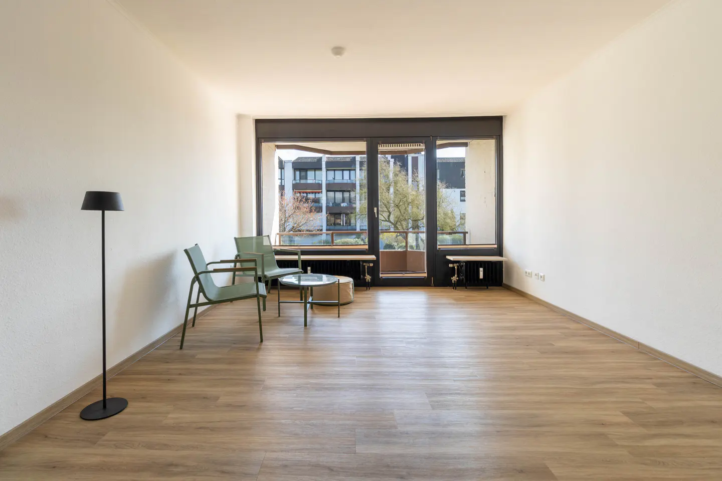 Bright, empty room with wood floors, white walls, and a large window. Two green chairs and a small table sit near the window. A black floor lamp stands in the corner.