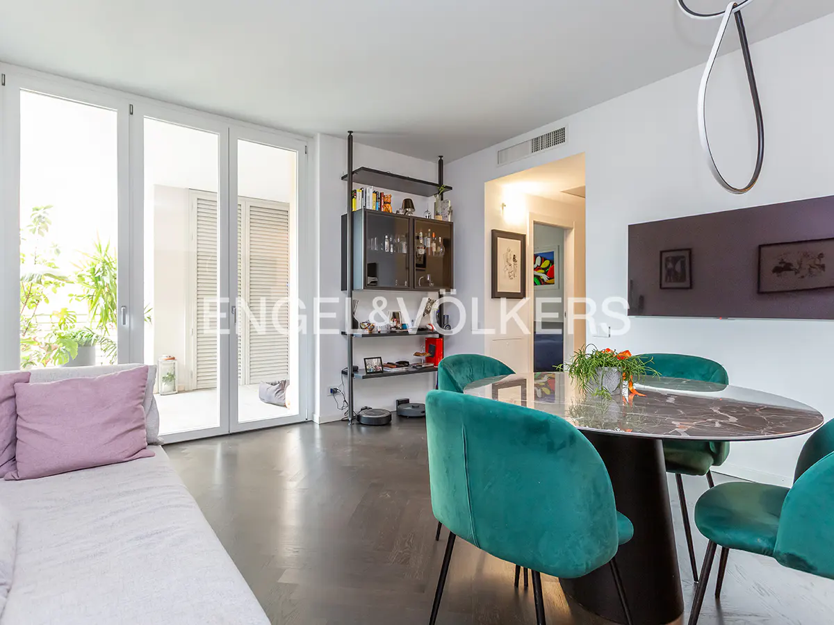 Bright living room with a white sofa, pink pillows, a round marble table with green velvet chairs, and a balcony with plants.