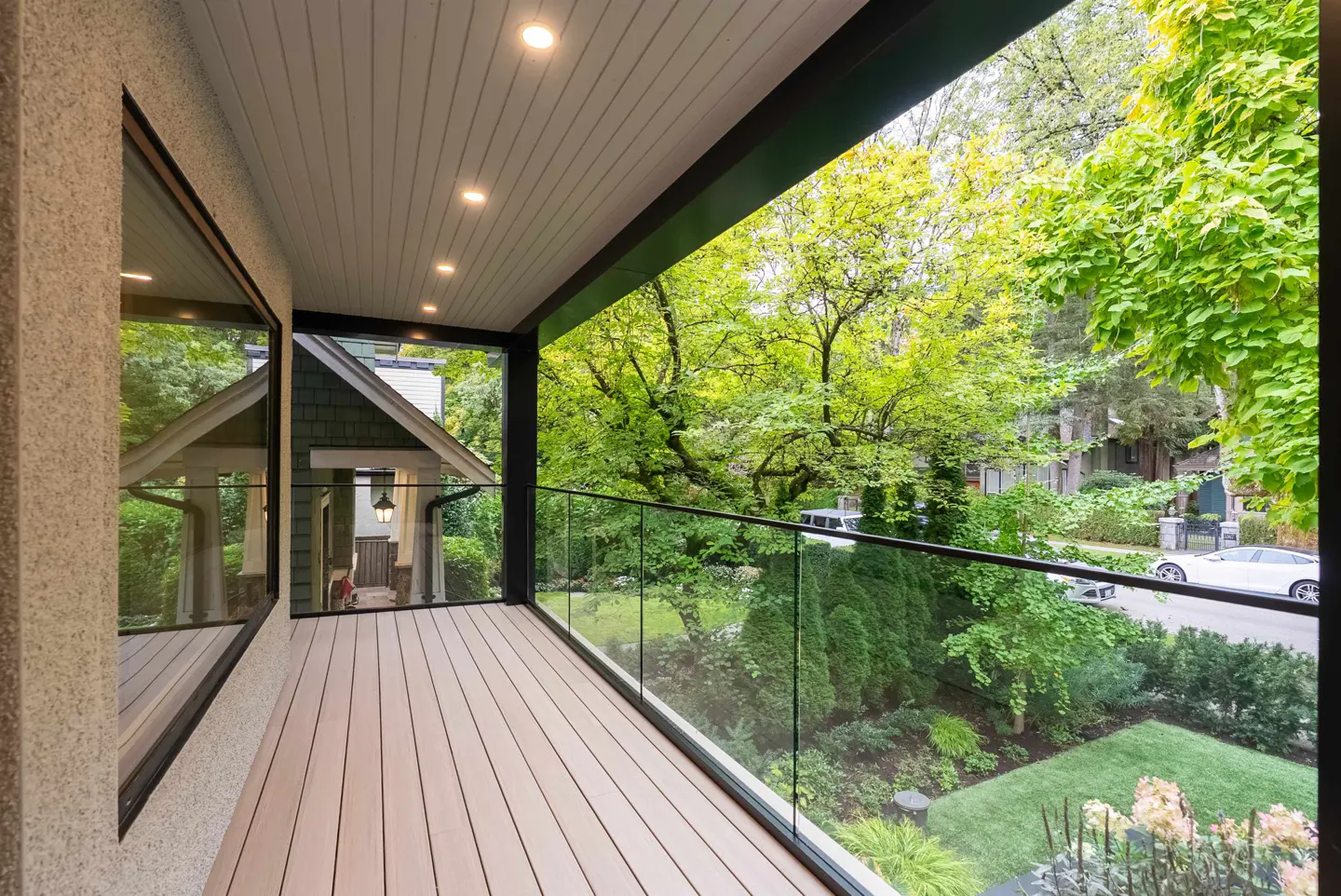 A modern balcony with wood flooring, glass railings, and a black frame overlooks a lush green neighborhood with trees and houses.