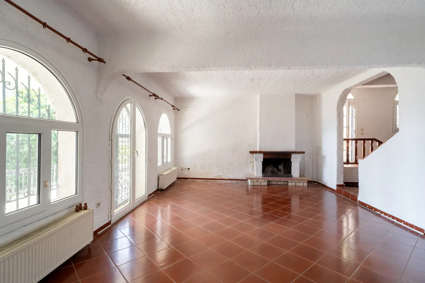 Bright, empty room with white walls, arched windows, and terracotta tile floor. A fireplace is on the far wall, and a staircase is visible through an archway.