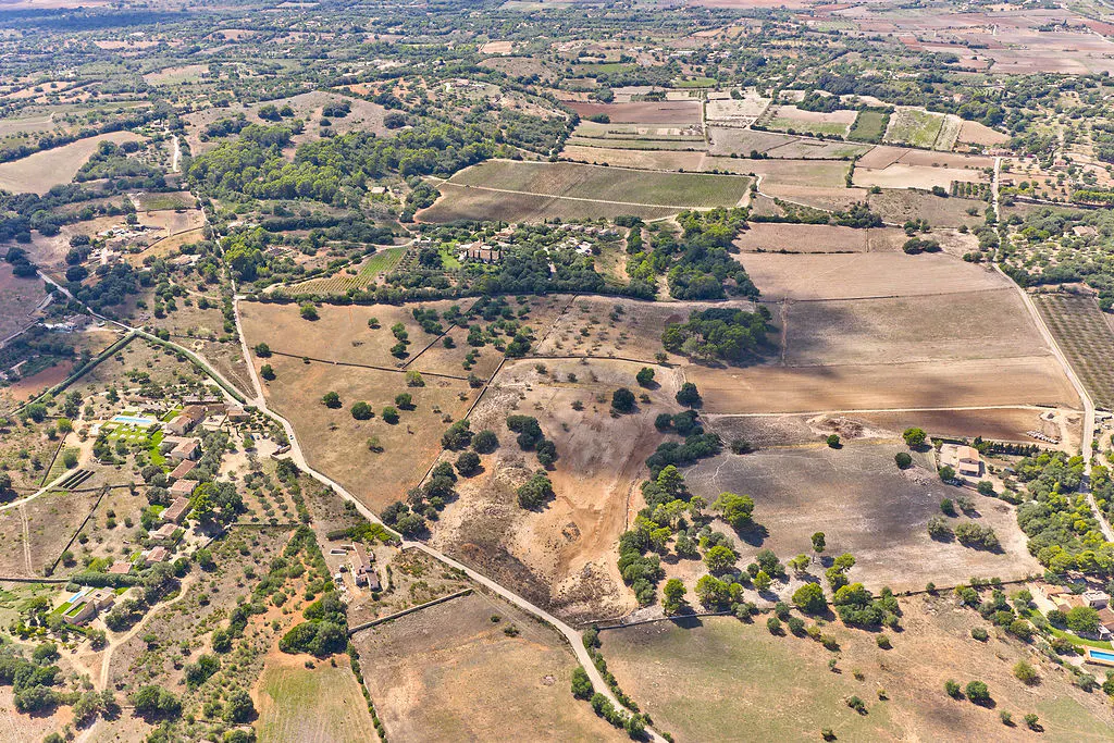 Aerial view of a rural landscape with fields, trees, and scattered buildings under a sunny sky.