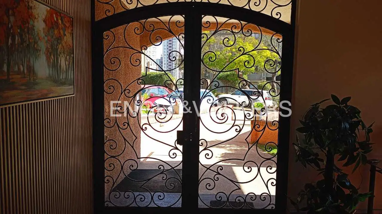 Ornate black wrought iron double doors offer a glimpse of a sunny street with cars and greenery. A painting hangs on the left wall.
