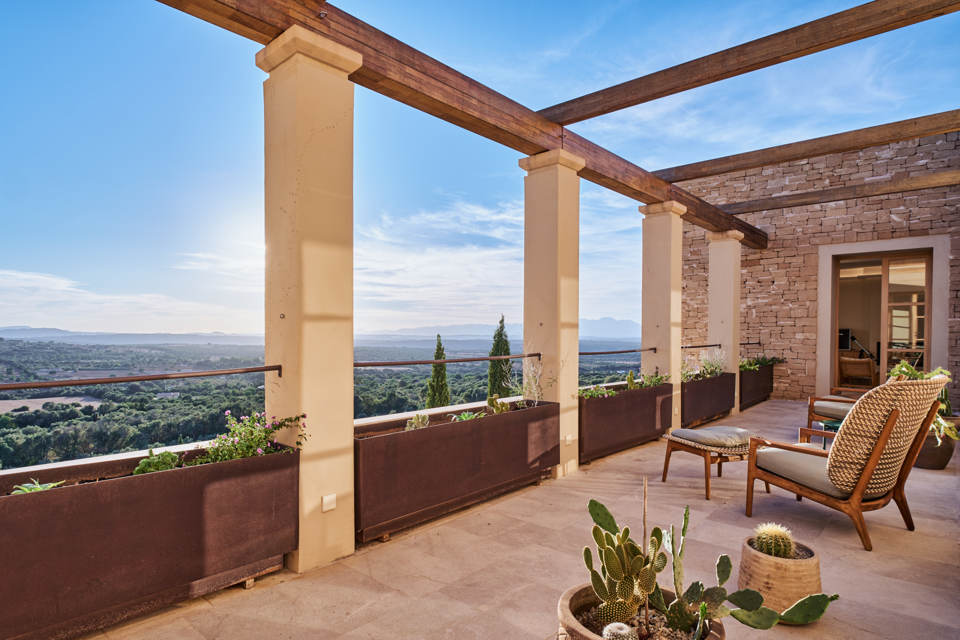 A sunny stone terrace with a wooden pergola, potted cacti, and lounge chairs overlooking a vast, scenic landscape. [AI-generated text]