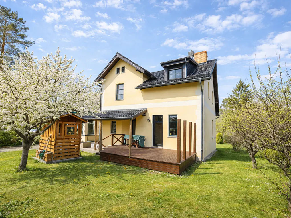 Yellow two-story house with a wooden deck, a small shed, and a blooming tree in a sunny green backyard.