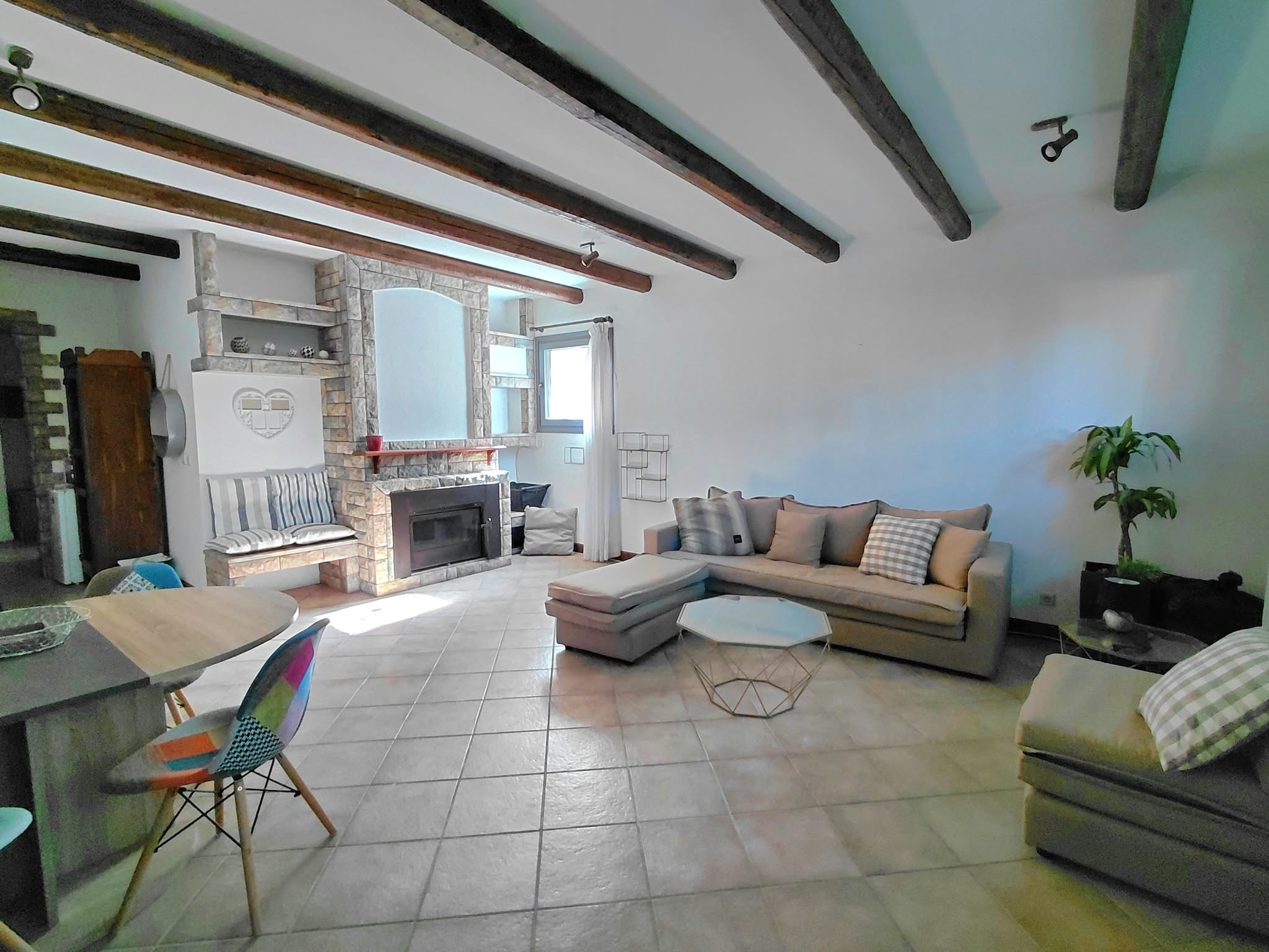 Spacious living room with tile floors, exposed wood ceiling beams, a stone fireplace, and a large beige sectional sofa. [AI-generated text]