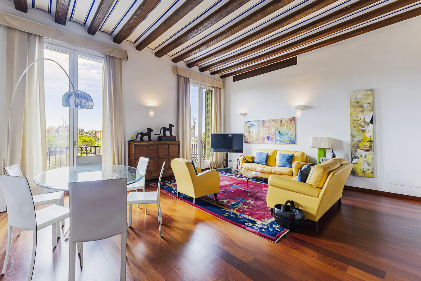 Bright living room with wood floors, yellow sofa and chairs, glass table, and exposed beam ceiling. Balcony view.
