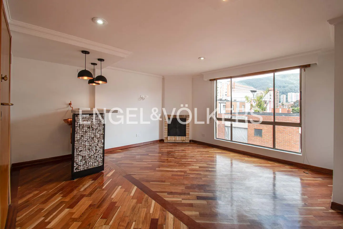 Modern living room with polished wood floors, a corner fireplace, a small bar area with pendant lights, and city views.