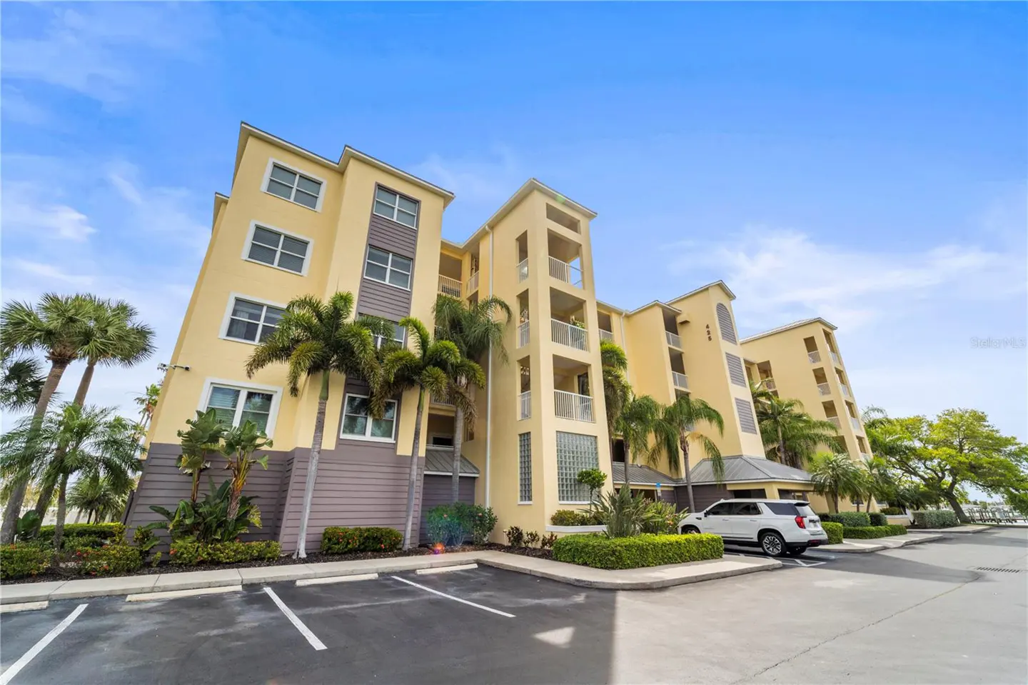 Exterior view of a yellow condo building with palm trees, a white SUV, and parking spaces under a blue sky.