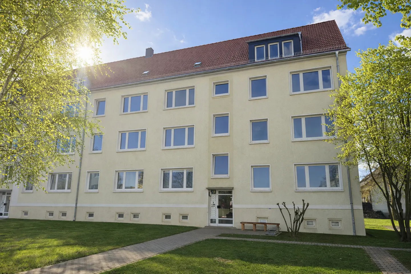 Three-story apartment building with a red roof and a light yellow facade, surrounded by green trees and grass.