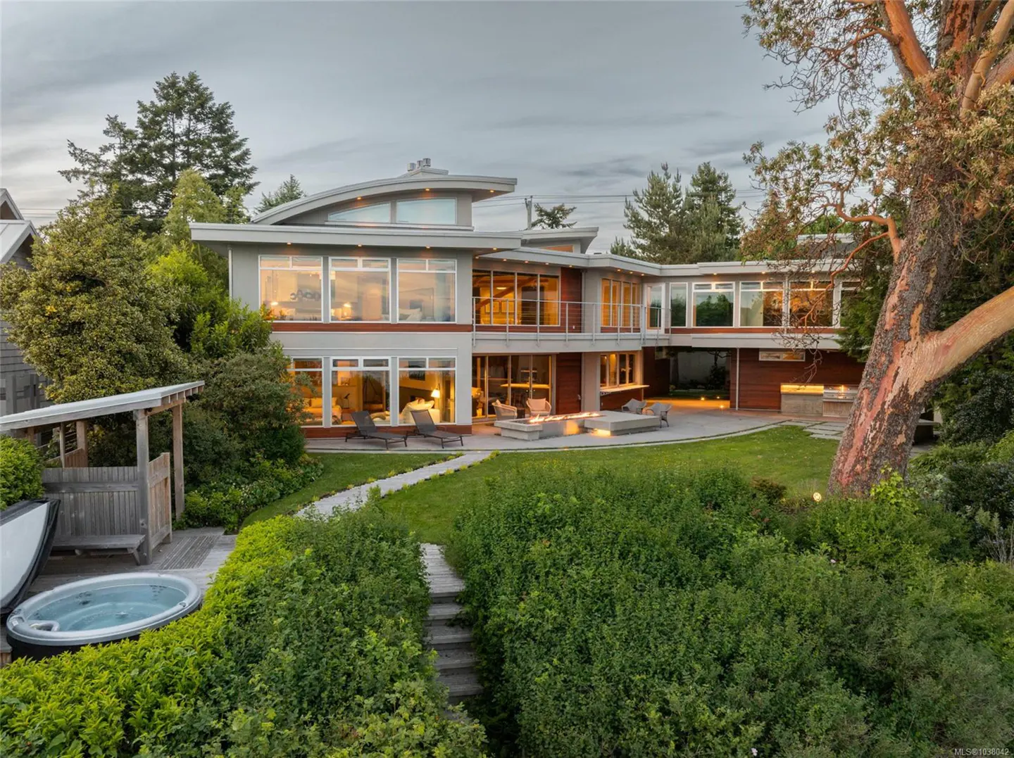 Two-story modern house with large windows, a green lawn, and a hot tub. A fire pit is on the patio.
