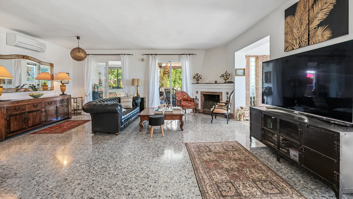 Bright living room with black leather sofa, wooden furniture, and a large TV. A fireplace and garden view add to the room's charm.