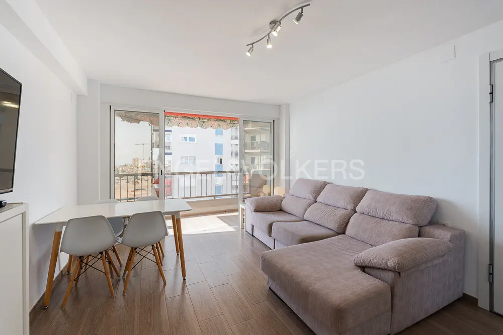 Bright living room with a beige sofa, white table and chairs, and a balcony overlooking a city.