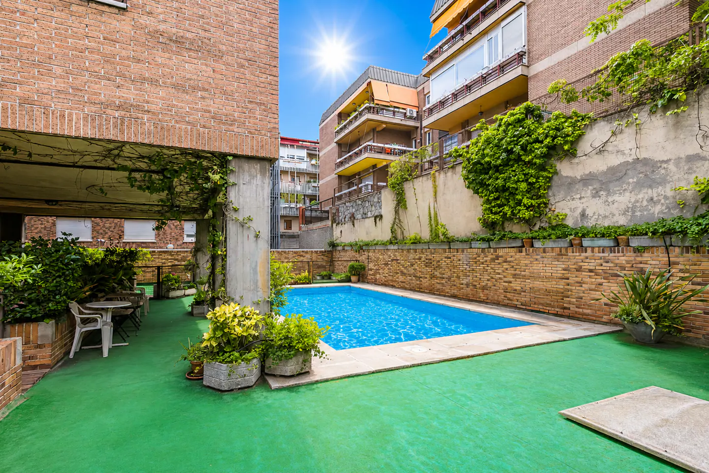Outdoor swimming pool with bright blue water, surrounded by a green patio, brick walls, and lush plants under a sunny sky.