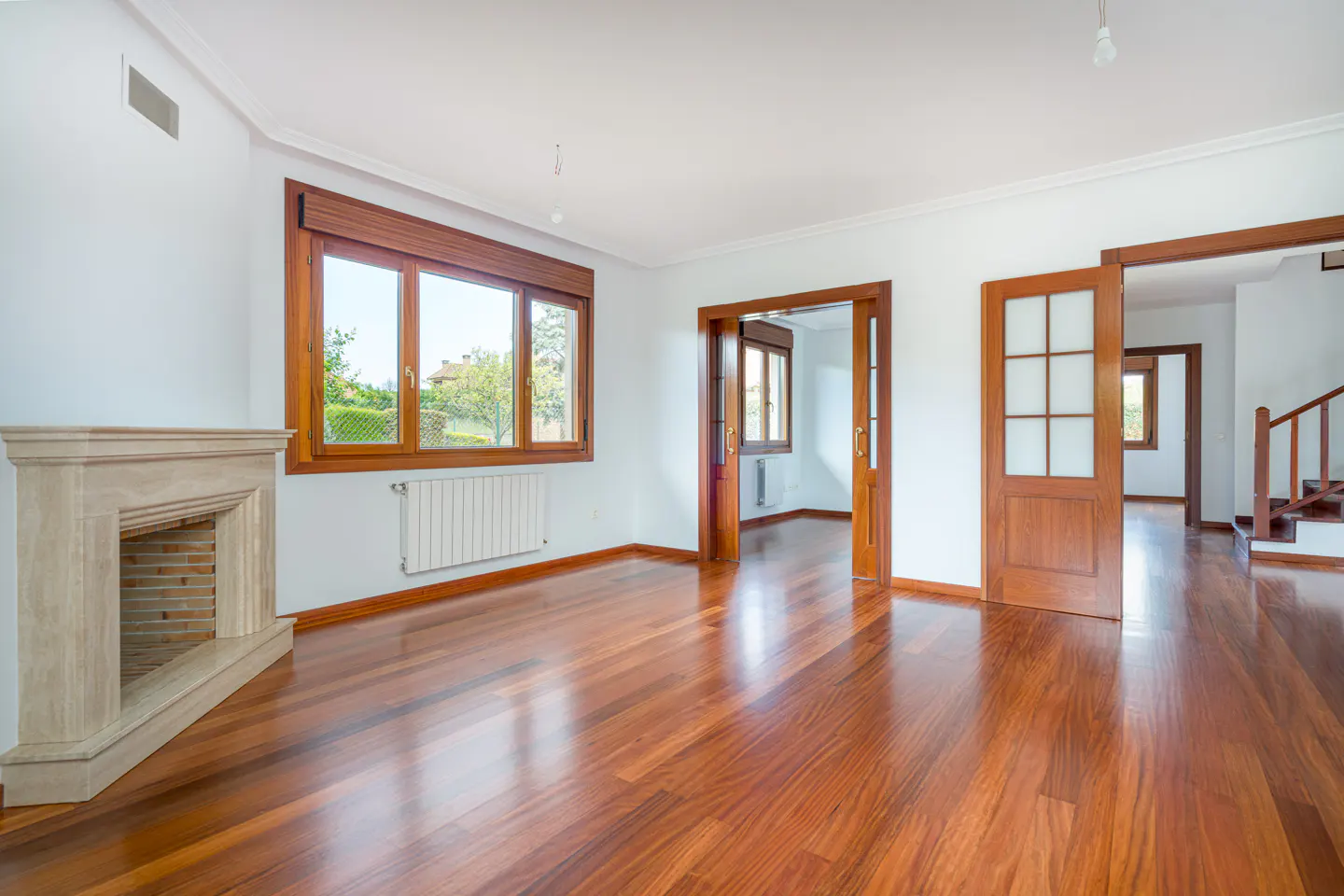 Empty living room with hardwood floors, white walls, a stone fireplace, and a large window with a view of greenery.