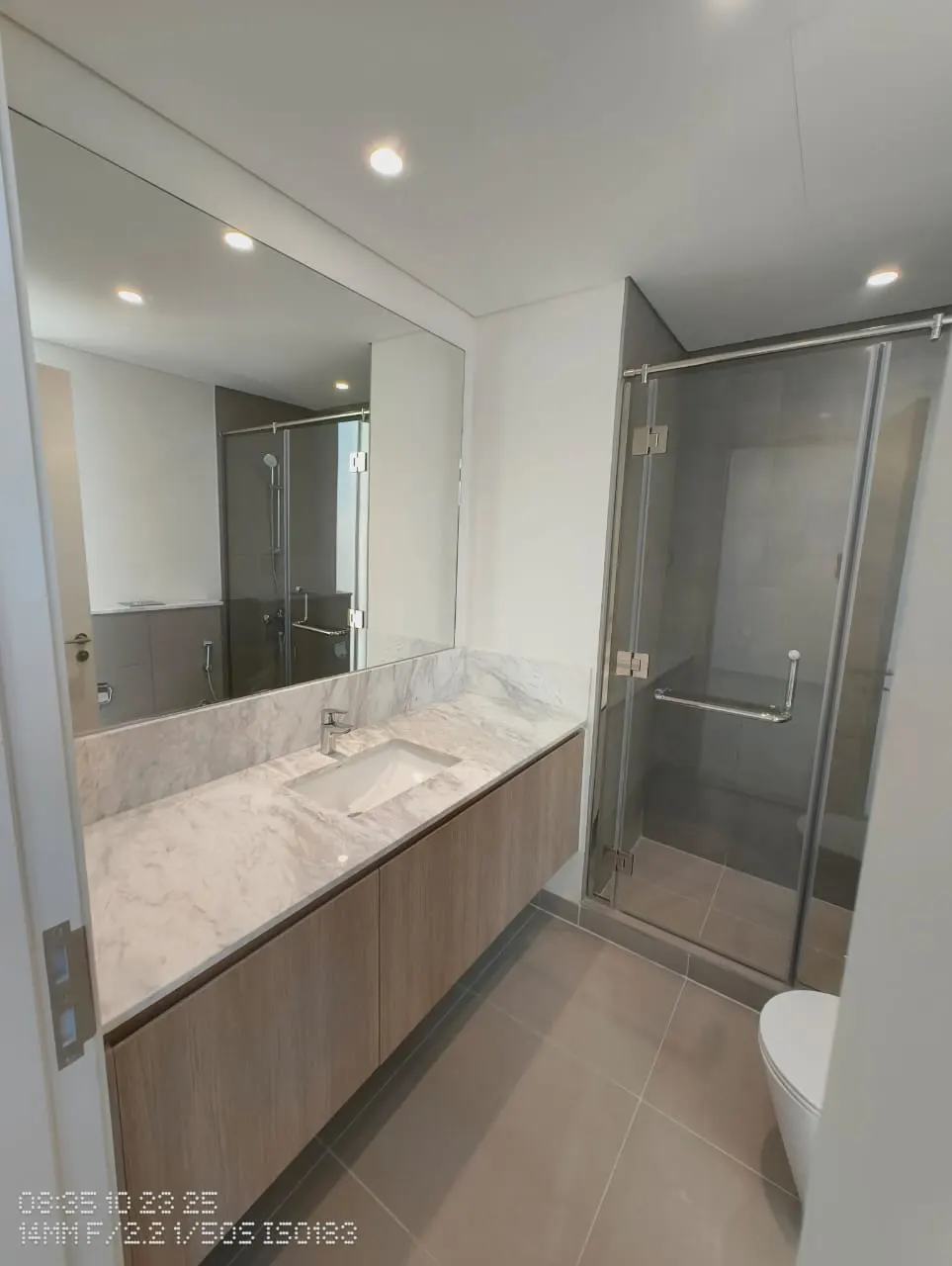 Modern bathroom with gray tile floor, marble countertop vanity, large mirror, and glass-enclosed shower. White toilet visible.