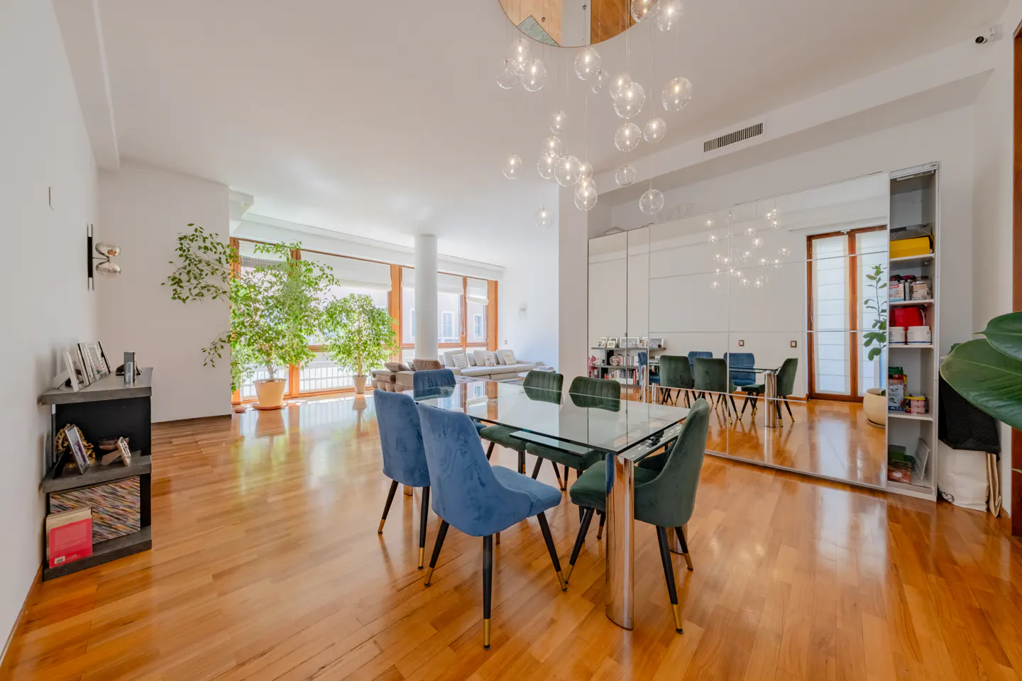 Bright, open dining area with a glass table, blue and green velvet chairs, and a modern bubble chandelier. Hardwood floors and large windows.