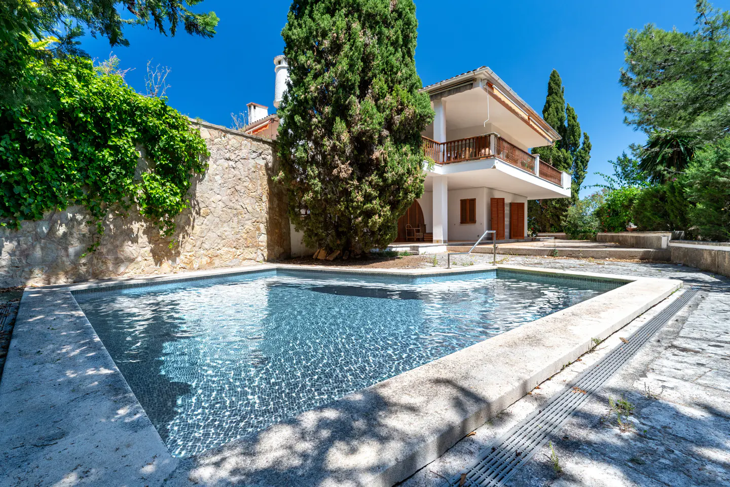 A two-story white house with a balcony overlooks a blue swimming pool on a sunny day.