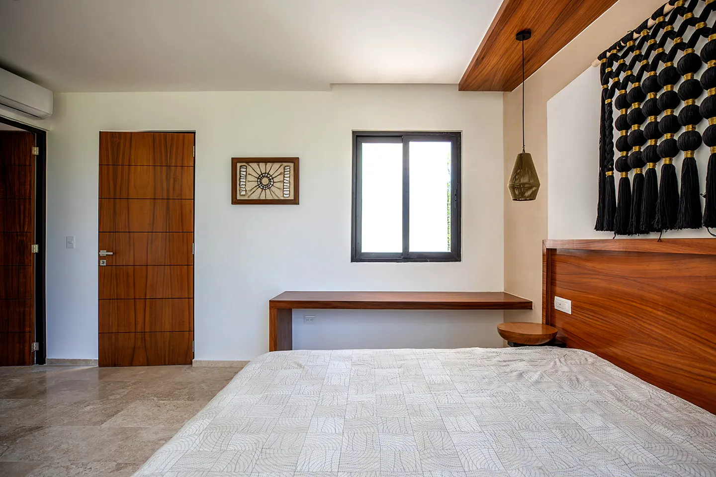 Bedroom with a wooden headboard, white patterned bedding, and a black tassel wall hanging. A desk sits under a window.