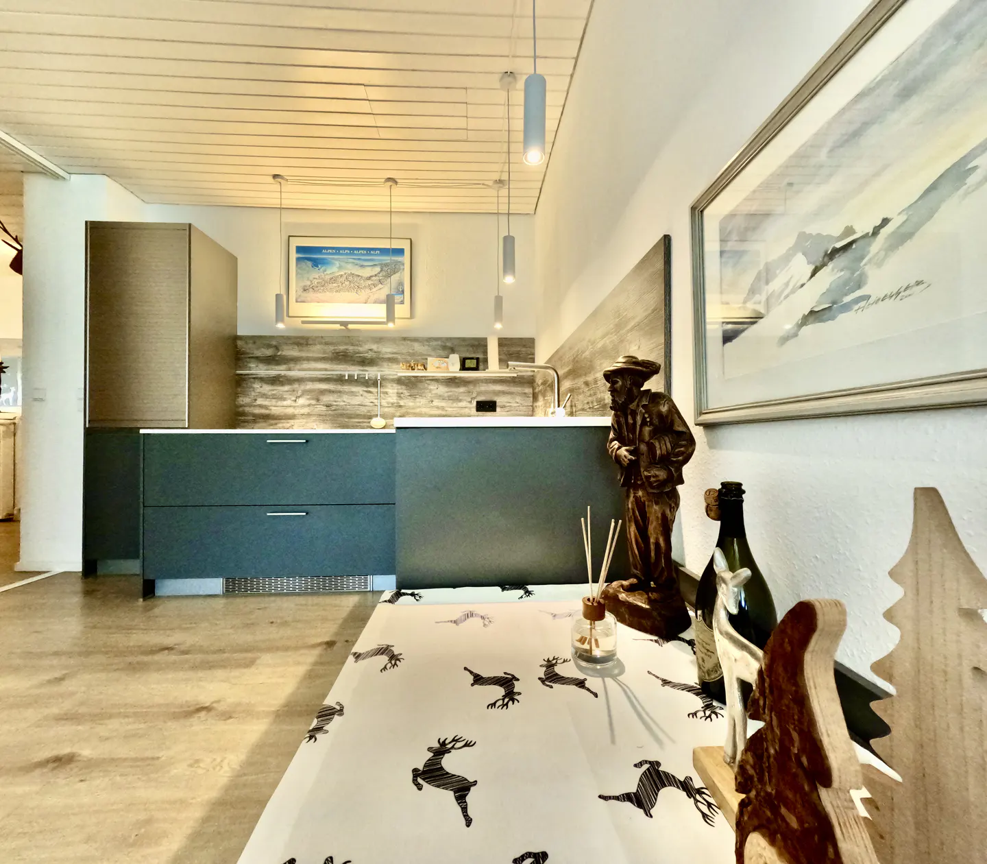 A modern kitchen with gray cabinets, wood floors, and a table with a reindeer-patterned tablecloth. A wooden statue and mountain painting add rustic charm.