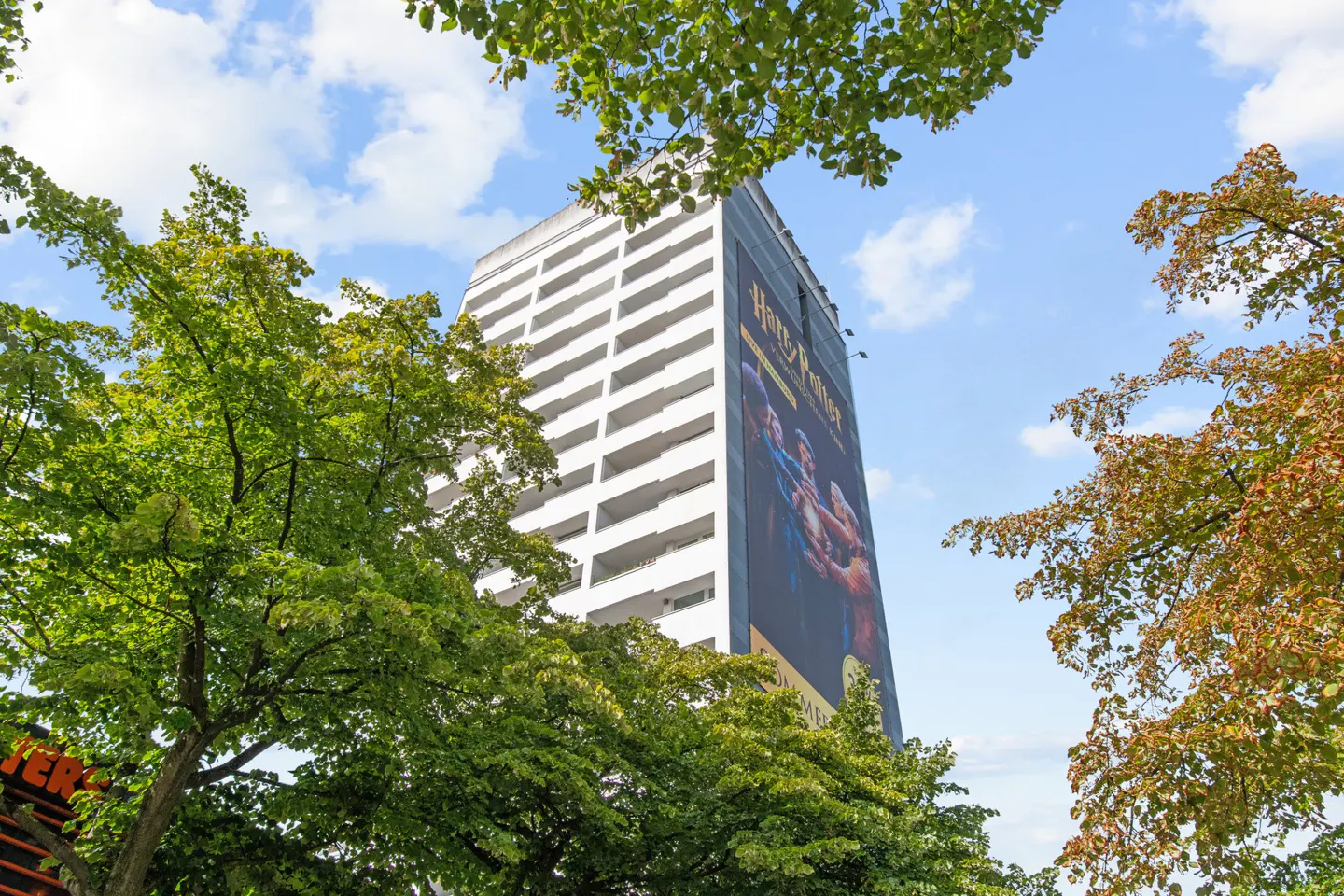 Low angle view of a tall, white apartment building with a large "Harry Potter" movie poster on its side, framed by green trees.