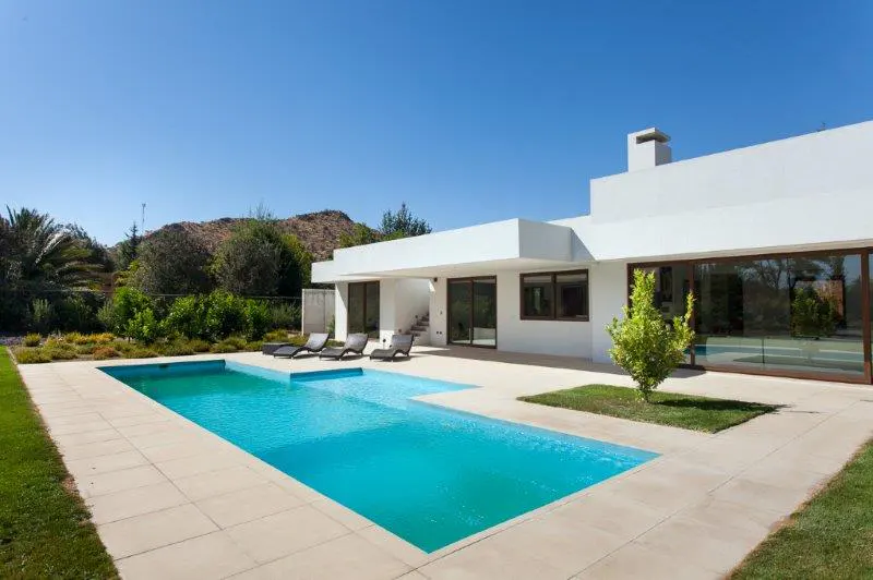 Modern white house with a turquoise pool, beige patio, and lounge chairs under a clear blue sky.